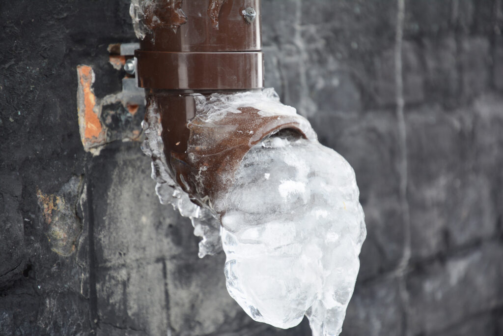 A plastic downspout, downpipe of a roof gutter system with ice and frozen water in the pipe near a house foundation in winter.